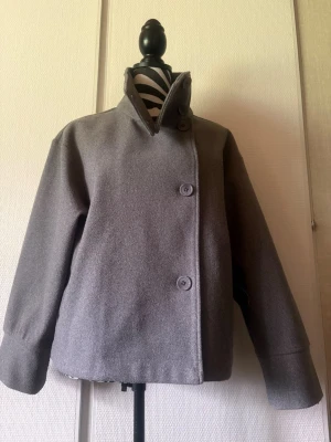 Grå ulljacka med hög krage - Stilren grå jacka i ullmix med hög krage och asymmetrisk knäppning framtill. Jackan har två stora knappar och vida ärmar för en modern look. Perfekt för dig som gillar minimalistisk och clean stil.