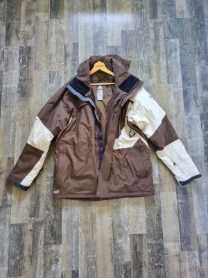Brun och beige vindjacka med huva från nike - Snygg vindjacka från Nike i brunt och beige med svarta detaljer. Jackan har huva, dragkedja och tryckknappar framtill samt två sidofickor. Tillverkad i slitstarkt syntetmaterial, perfekt för blåsiga dagar.