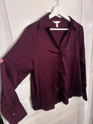 Vinröd blus med knappar från H&M - Säljer en vinröd blus från H&M med klassisk krage och knappar framtill. Blusen har lång ärm med bred manschett och är gjord i ett glansigt, mjukt material som ger en lyxig känsla. Perfekt för dig som gillar stilrena plagg med en twist.