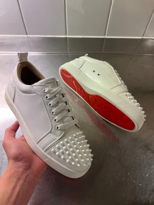 Vita sneakers med nitar från Louboutin - Snygga vita sneakers i läder från Christian Louboutin med ikoniska röda sulor och coola vita nitar på tån. Skorna har klassisk låg modell, vita skosnören och en stilren design som sticker ut. Perfekta för dig som vill ha något unikt och trendigt.
