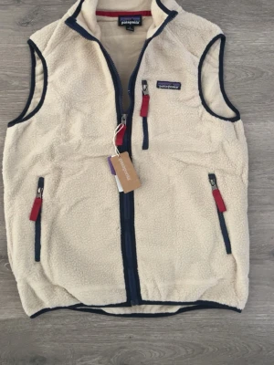 Beige fleeceväst från Patagonia - Säljer en beige fleeceväst från Patagonia med marinblå detaljer och röda dragkedjor. Västen har hög krage, tre fickor med dragkedja och är perfekt för lager-på-lager. Klassisk och stilren design som funkar året runt, aldrig använd, passade inte mig