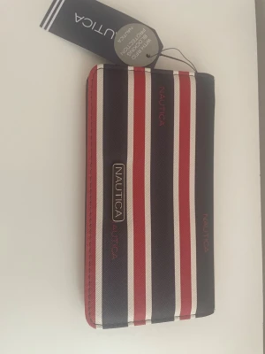 Randig plånbok från Nautica - Snygg plånbok från Nautica med marinblå, röda och vita ränder. Plånboken har ett rektangulärt format och är tillverkad i slitstarkt syntetmaterial. Märkets logga syns tydligt på framsidan i metall och tryck. Perfekt för dig som gillar klassisk och sportig stil.