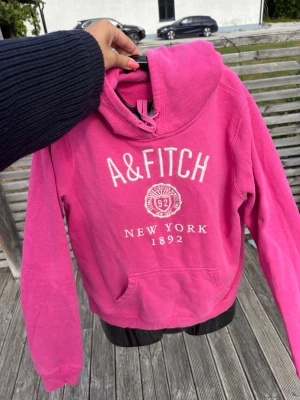 Rosa hoodie från A&Fitch - Snygg rosa hoodie från A&Fitch med tryck framtill där det står 'A&FITCH New York 1892'. Hoodien har huva med dragsko och en klassisk känguruficka. Perfekt för en avslappnad och trendig look.