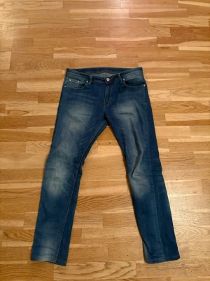 Blå jeans från Nudie Jeans - Säljer ett par klassiska blå jeans från Nudie Jeans med slitningar och tvättade detaljer. Modellen har fem fickor, slimfit och raka ben. Perfekta för dig som gillar en avslappnad men snygg stil.
