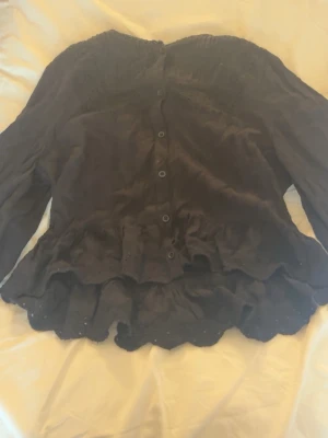 Svart blus med volang från Zara - Snygg svart blus från Zara med knappar framtill och volangdetaljer längs nederkanten. Blusen har långärmad design och fina spetsdetaljer vid axlar och halsringning. Tillverkad i ett lätt och luftigt tyg som ger en skön känsla.
