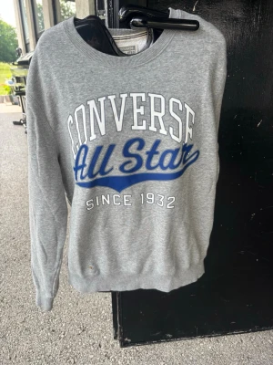 Grå sweatshirt från Converse - Snygg grå sweatshirt från Converse med stort tryck 'Converse All Star Since 1932' i vitt och blått på bröstet. Klassisk rund halsringning och mjukt material som passar perfekt till jeans eller joggers. Enkel och stilren design för en avslappnad look.