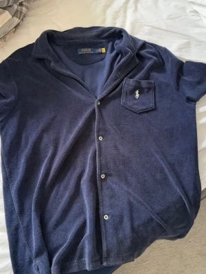 Marinblå frottéskjorta Polo Ralph Lauren - Säljer en marinblå skjorta från Polo Ralph Lauren i mjuk frotté. Skjortan har korta ärmar, klassisk krage, knappar framtill och en bröstficka med broderad logga. Perfekt för en avslappnad och cool stil.