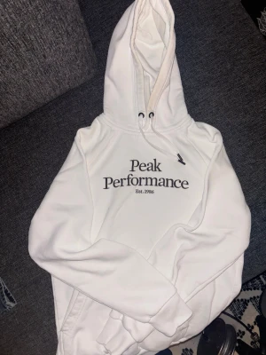 Vit hoodie från Peak Performance - Säljer en vit hoodie från Peak Performance med svart logga och text på bröstet. Hoodien har huva med dragsko och en klassisk, avslappnad passform. Perfekt för dig som gillar stilrena och bekväma plagg. Materialet känns mjukt och skönt mot huden.