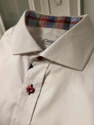 Oscar of Sweden slim fit two fold vit skjorta röda detaljer, storlek 41 16 - Klassisk vit skjorta från Oscar of Sweden med röda knappar och rutigt mönster på insidan av kragen. Skjortan har lång ärm, normal passform och är tillverkad i bomull. Perfekt för dig som vill ha en stilren look med en twist.