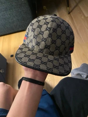 Gucci keps med GG-mönster - Säljer en snygg keps från Gucci med klassiskt GG-monogram i beige och svart. Kepsen har böjd skärm och detaljer med röd och marinblå rand på sidorna. Justerbar rem bak och tillverkad i canvasmaterial. Perfekt för dig som vill ha en lyxig och trendig accessoar.