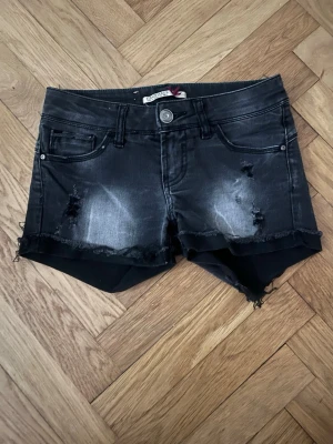Svarta lågmidjade korta jeansshorts - Säljer ett par svarta jeansshorts med slitna detaljer och råa kanter. Shortsen har fem fickor, knappgylf och en cool, lite tvättad look. Perfekta för dig som gillar en edgy stil och vill ha något bekvämt till sommaren💕