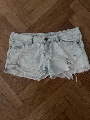 Ljusblå korta lågmidjade jeansshorts - Säljer ett par ljusblå, vintage jeansshorts med råa, fransiga kanter och slitna detaljer framtill. Klassisk femficksmodell med knapp och dragkedja. Jättesöta och perfekta för sommaren💕