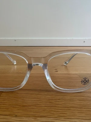 Transparenta solglasögon med blommig detalj - Säljer ett par stilrena solglasögon med transparent båge och en liten blommig detalj på ena linsen. Rektangulär form och modern look som passar till många outfits. Lätta och bekväma att bära, perfekta för dig som gillar minimalistisk stil.