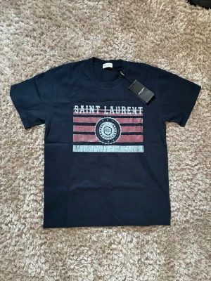 Svart Saint Laurent t-shirt - Snygg svart t-shirt från Saint Laurent med tryck i vitt och rött framtill. T-shirten har klassisk passform, rund halsringning och korta ärmar. Materialet är mjuk bomull och trycket har en vintagekänsla med Saint Laurent-logga.