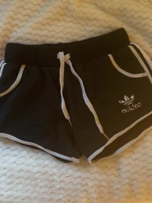 Vintage svarta adidas shorts - Svarta shorts från Adidas med vita kanter och snörning i midjan. Klassisk Adidas-logga på ena benet och bekväm passform. 