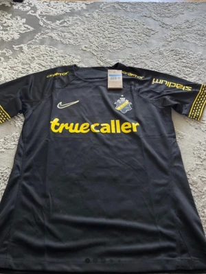 AIK matchtröja Nike svart/gul - Snygg svart AIK matchtröja från Nike med gula detaljer och tryck från Truecaller, SVEA och Stadium. AIK-emblemet på bröstet och årtalen 1934-2024 i nacken. Perfekt för dig som vill stötta laget på match eller träning.