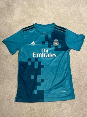 Real Madrid Ronaldo T-shirt - Snygg turkosblå Real Madrid matchtröja från Adidas med Ronaldo och nummer 7 på ryggen. Tröjan har korta ärmar, svarta detaljer på axlarna och grafiskt mönster framtill. Tillverkad i lätt och ventilerande material, perfekt för fotbollsträning.