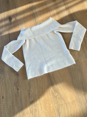 Vit off-shoulder stickad tröja - Säljer en vit stickad tröja med bred off-shoulder krage och ribbade detaljer vid ärmslut och nederkant. Tröjan har lång ärm och är gjord i ett mjukt material. Perfekt för dig som gillar en stilren och trendig look.