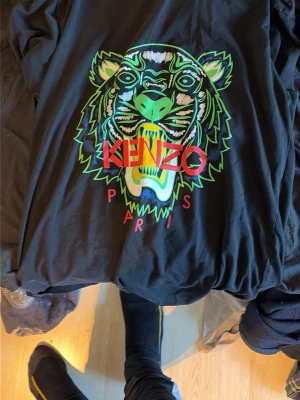 Svart Kenzo t-shirt med tigertryck - Cool svart t-shirt från Kenzo med stort färgglatt tigertryck och röd Kenzo Paris-text på bröstet. T-shirten har klassisk passform och är tillverkad i mjuk bomull. Perfekt statement-plagg för dig som vill sticka ut.