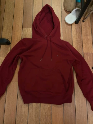 Vinröd hoodie från Tommy Hilfiger - Snygg vinröd hoodie från Tommy Hilfiger med klassisk huva och dragsnören. Hoodien har en stor magficka och diskret Hilfiger-logga på bröstet. Tillverkad i mjukt bomullsmaterial som känns skönt mot huden. Perfekt för en chill och stilren look. Priset kan absolut diskuteras. Nypris ca 1000 kr
