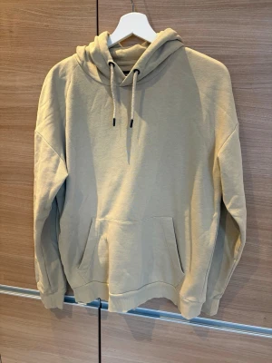Beige hoodie med huva - Säljer en beige hoodie med stor känguruficka framtill och justerbar huva med snören. Hoodien har ribbade muddar vid ärmslut och nederkant. Perfekt för en chill och avslappnad stil. Materialet känns mjukt och skönt, troligen bomullsblandning.