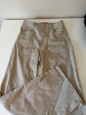 Beige raka jeans med hög midja - Snygga beige jeans med raka ben och hög midja. Klassisk femficksmodell med bälteshällor och knappgylf. Materialet är mjukt och bekvämt, perfekt för en avslappnad stil. Passar till det mesta och ger en clean look.