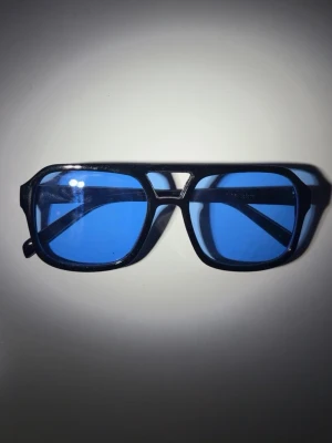 Svarta solglasögon med blåa glas - Snygga solglasögon med svart, kantig båge och blåa tonade glas. Glasögonen har en retroinspirerad design med breda skalmar och markerad näsbrygga. Perfekta för dig som vill sticka ut med en cool accessoar.