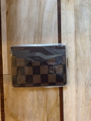 Brun rutmönstrad plånbok Louis Vuitton - Snygg plånbok från Louis Vuitton i klassiskt brunt damier-mönster. Tillverkad i läder med rutigt tryck och diskret logotyp på framsidan. Smidig och kompakt modell med plats för kort och sedlar. Perfekt accessoar för dig som gillar lyxig och tidlös design.