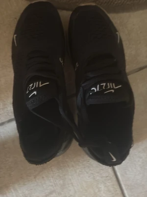 Nike Air Max 270 svart sneakers - Svarta Nike Air Max 270 sneakers med mesh-överdel och tydlig Air Max-dämpning i hälen. Skorna har klassisk Nike-logga på tungan och sidan samt snörning. Snygg och sportig design som passar perfekt till streetwear.