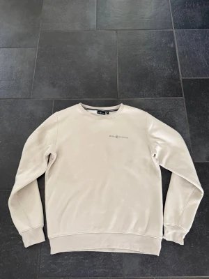 Sail Racing  - Beige tjocktröja från Sail Racing med diskret logga på bröstet. Tröjan har rund halsringning, långa ärmar och ribbade muddar vid ärmslut och nederkant. Perfekt för en clean och stilren look.