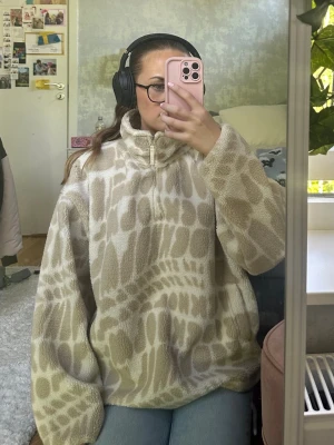 Beige och vit fleece-tröja med mönster - Mysig beige och vit fleece-tröja med halv dragkedja och högt krage. Tröjan har ett unikt, abstrakt mönster och är oversized för en avslappnad look. Perfekt för kalla dagar och enkel att styla med jeans eller leggings.