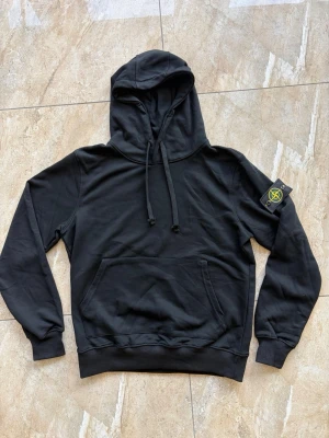 Svart Stone Island Hoodie - Snygg svart hoodie från Stone Island med klassisk patch på vänster ärm. Hoodien har dragsko i huvan och en stor magficka framtill. Tillverkad i mjuk bomull som känns skön mot huden. Perfekt för dig som gillar streetwear och vill ha en stilren look och passar i storlek L