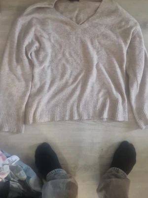 Beige stickad tröja från Amisu - Mysig beige stickad tröja från Amisu med v-ringning och långa ärmar. Tröjan har ribbade muddar vid ärmslut och nederkant, vilket ger en snygg och avslappnad look. Perfekt att matcha med jeans eller kjol för en enkel stil.