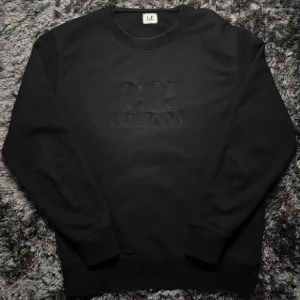 C.P. Company Sweatshirt - Klassisk svart C.P. Company-sweatshirt med broderad logga framtill. Storlek L, men passar även M beroende på önskad passform. Stilren, lättmatchad och perfekt till vardags/streetwear. QR-kod finns som äkthetsbevis. Skick enligt bilder.