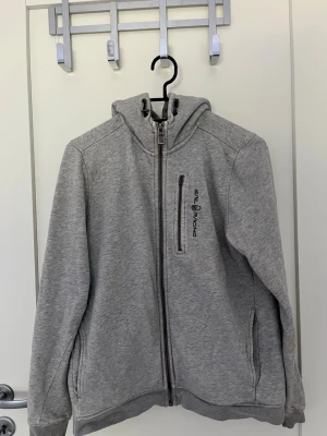Grå hoodie från Sail Racing - Snygg grå hoodie från Sail Racing med dragkedja framtill, bröstficka med logga och två sidofickor. Mjuk bomullsmix och klassisk passform med huva. Perfekt för en avslappnad och sportig stil.