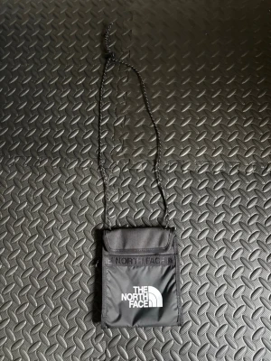 Svart axelväska The North Face - Snygg svart axelväska från The North Face med justerbar tunn axelrem. Väskan har en fyrkantig form, lockstängning och tydlig logga framtill. Tillverkad i slitstark nylon, perfekt för att bära det viktigaste på stan eller resa.