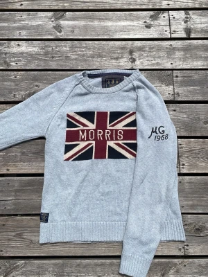 Grå stickad tröja Morris - Ljusblå stickad tröja från Morris med Union Jack-flagga och texten 'MORRIS' på bröstet. På ärmen står det 'MG 1968' broderat. Tröjan har rund halsringning och ribbade muddar. Perfekt för dig som gillar klassisk brittisk stil.