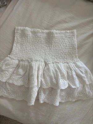 Vit volangkjol med broderade detaljer - Supersöt vit kortkjol med bred smock i midjan och dubbla volanger. Kjolen har broderade blommönster och hålbroderier som ger en romantisk känsla. Perfekt för varma dagar och lätt att matcha med crop tops eller linnen.