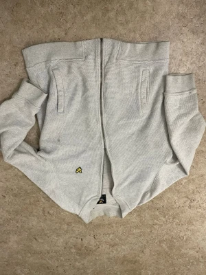 Ljusgrå ziptröja från Lyle & Scott - Ljusgrå ziptröja från Lyle & Scott med gul logga på vänster sida. Tröjan har två fickor framtill, ribbade muddar och är tillverkad i ett strukturerat bomullstyg. Perfekt för en avslappnad och stilren look.