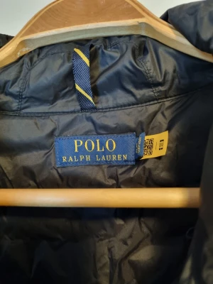 Ralph Lauren väst - Snygg svart dunväst från Polo Ralph Lauren med quiltad design och hög krage. Västen har två dragkedjeförsedda fickor och en diskret röd logotyp på bröstet. Perfekt för lager-på-lager och håller dig varm under kyliga dagar.