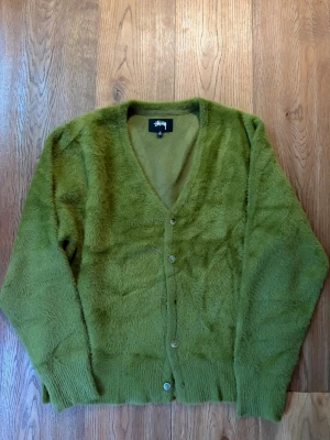 Grön shag kofta från Stussy - Säljer en grön, fluffig kofta från Stussy med v-ringning och knäppning framtill. Koftan har långa ärmar och ribbade muddar vid ärmslut och nederkant. Materialet är mjukt och ger en riktigt cozy känsla.