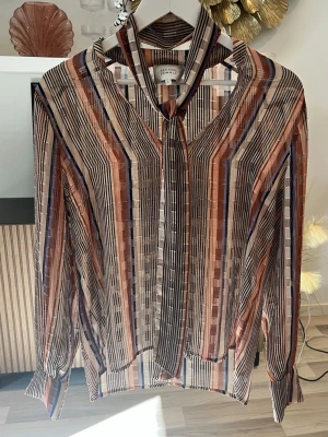 Randig blus med knytband från Second Female - Snygg transparent blus från Second Female med vertikala ränder i brunt, beige, svart och lila i storlek S. Blusen har knytband i halsen och långa ärmar med knapp vid ärmslut. Perfekt för dig som gillar trendiga mönster och lätta material. Axelbredd: 43cm. Byst: 50cm. Längd: 70cm. 