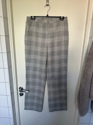 Grå rutiga kostymbyxor storlek 40 - Snygga grå kostymbyxor med klassiskt rutigt mönster och raka ben. Byxorna har hällor för skärp och stängs framtill. Perfekta för en stilren och trendig look. Materialet känns mjukt och följsamt.