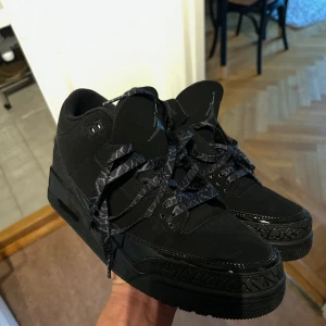 Svarta Air Jordan 3 sneakers - Säljer ett par helsvarta Air Jordan 3 sneakers med klassisk siluett. Skorna har detaljer i mocka och läder, samt mönstrade svarta skosnören. Ikoniska Jumpman-loggan syns på plösen och hälpartiet. Perfekta för dig som gillar stilrena och sportiga sneakers.