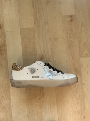Golden Goose vita sneakers med glitter - Snygga vita sneakers från Golden Goose med svart snörning, silverfärgad stjärna på sidan och guldglittrig häl. Skorna har en vintage-look med slitna detaljer. Nypris ca 5000kr