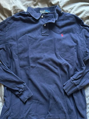 Marinblå långärmad piké från Polo Ralph Lauren - Klassisk marinblå långärmad pikétröja från Polo Ralph Lauren med röd broderad logga på bröstet. Skjortan har krage, knappar vid halsen och är gjord i mjuk bomull. Perfekt för en avslappnad men ändå stilren look.