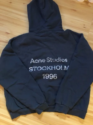 Svart hoodie Acne Studios Stockholm - Svart hoodie från Acne Studios med tryck på ryggen där det står 'Acne Studios STOCKHOLM 1996' i vitt. Klassisk modell med huva och muddar vid ärmslut och nederkant. Perfekt för en avslappnad och trendig streetwear-look.