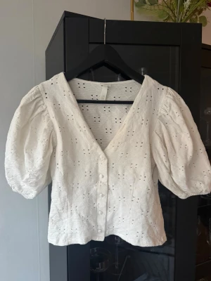 Vit blus med puffärm och broderi - Superfin vit blus med v-ringning, puffiga korta ärmar och broderade hålmönster. Knappar framtill och lätt struktur i tyget ger en romantisk känsla. Perfekt att styla med jeans eller kjol.