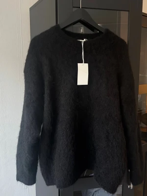 Svart fluffig stickad tröja NAKD - Mysig svart stickad tröja med fluffig yta och rund hals. Tröjan har långa ärmar och en avslappnad passform, perfekt för lager på lager. Materialet ser ut att vara en mjuk alpacka- eller ullblandning. Enkel och stilren design utan synliga detaljer. aldrig använd 