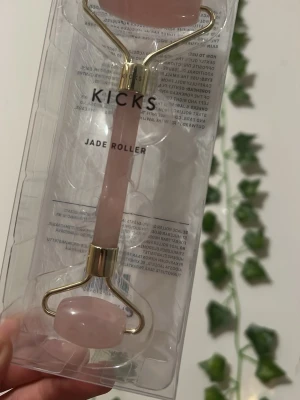 Jade roller från Kicks i rosa sten - En stilren jade roller från Kicks med handtag och rullar i ljusrosa sten och detaljer i guldmetall. Perfekt för ansiktsmassage och hudvård. Ger en lyxig känsla och är enkel att använda i din dagliga rutin.